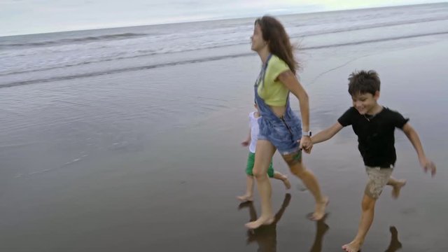 Follow shot of happy mother or sister and two little boys running barefoot along ocean coast when having fun at the beach during summer vacation