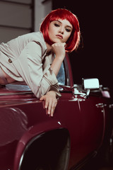 Fototapeta na wymiar attractive young woman in red wig and trench coat looking at camera while leaning at classic car