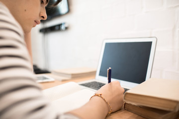 Attractive young male sitting at home at desk with laptop and making notes while reading book. 