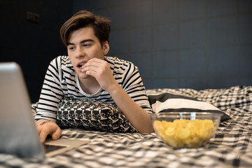 Young man  lying on bed and eating chips concentrated on using laptop computer.