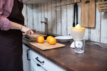 Teenager in apron standing in kitchen and cutting ripe oranges with knife