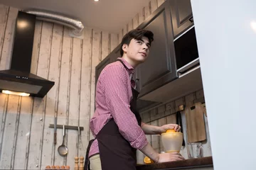Foto op Plexiglas Teenager in apron making orange juice and looking away while standing in kitchen  © Seventyfour