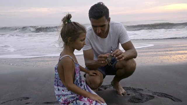 Loving father showing and telling about crab to little daughter while sitting with on sand at ocean beach