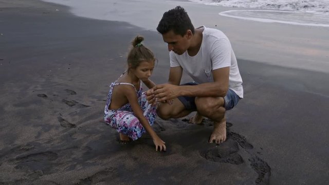Little girl playing with wet sand on ocean beach and speaking with dad while he showing crab to her