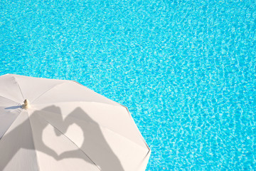 Shadows of hands forming a heart on a white parasol, blue swimming pool water background, summer concept © Delphotostock