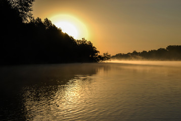 A  view of a yellow sunrise under the calm river. The sun lifts over the trees and sunlight is very beautiful thourgh the mist on the river. Orihivka river, Ukraine. Concept - mysterious nature.