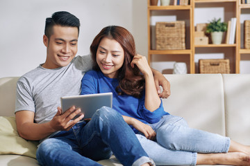 Beautiful Asian young couple embracing and chilling on couch at home while watching tablet together