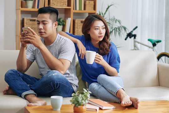 Asian Couple Sitting On Couch And Man Surfing Smartphone While Woman Looking Bored And lonely