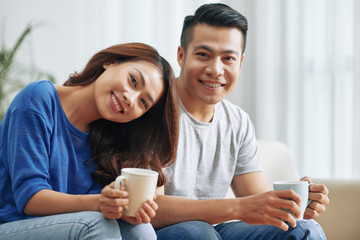 Content Asian man and woman holding coffee cups and smiling happily at camera chilling on sofa