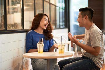 Asian young couple chilling in cafe having drinks in cups and laughing happily while chatting