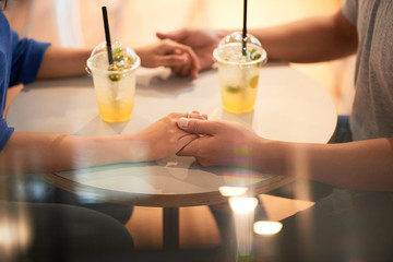 Faceless side view of couple sitting with drinks at table and holding hands on top in lights
