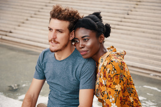 Tender black woman leaning on man shoulder looking away in dream and love while sitting on street