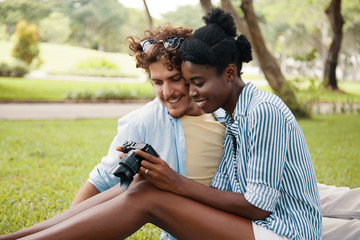 Young black woman and Caucasian man sitting on meadow in park and watching photo camera in leisure