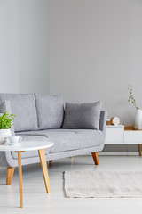 Real photo of a grey sofa standing between a small table and a shelf in a simple, grey living room interior