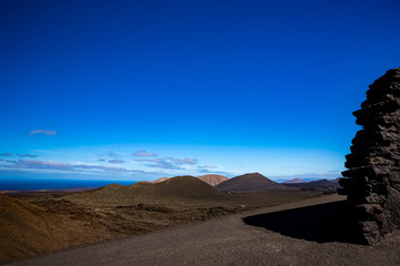 Gravel road in the vast emptiness and loneliness of the Lanzarote black frozen lava volcanic desert and few sleeping volcanoes in the background. Stone wall in the right-hand side of the image