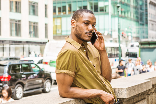 Young African American Man with beard, wearing green short sleeve shirt, standing against half wall, back facing busy street in Midtown of Manhattan, New York, relaxing, talking on cell phone
