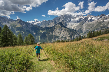 Fototapeta na wymiar A young boy hiking on a mountain path on Italian alps. A male with hat and holds trekking poles in her hands. Relaxing alone in a beautiful summer sunny day. A kid enjoying outdoor sport.
