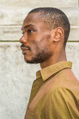 Close up, head short of Young African American Man with beard in New York, wearing green shirt,  collar unbuttoned, standing against white marble wall, seriously looking forward. Left Side View.