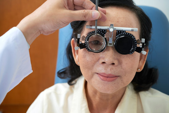 Crop hand of doctor setting special examining trial lens eyeglasses on face of adult Asian woman in ophthalmology cabinet