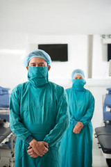 Fototapeta na wymiar Asian man in mask and coat of surgeon standing in operating room with female colleague on background