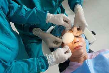 Crop shot from above of careful surgeons applying patch with tape on eye of mature Asian patient...