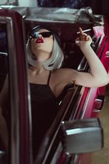 Fototapeta na wymiar stylish blonde woman in sunglasses and beret smoking cigarette while sitting in retro car
