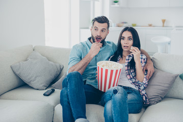Portrait of stressed astonished spouses with wide open mouth eyes eating snack having bucket of popcorn enjoying horror film sitting on couch indoor