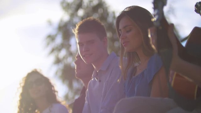 Attractive young friends hipsters singing songs with a guitar at summer sunset outdoors