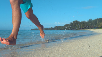 LOW ANGLE: Unknown young man runs in the shallow sea along the exotic beach.