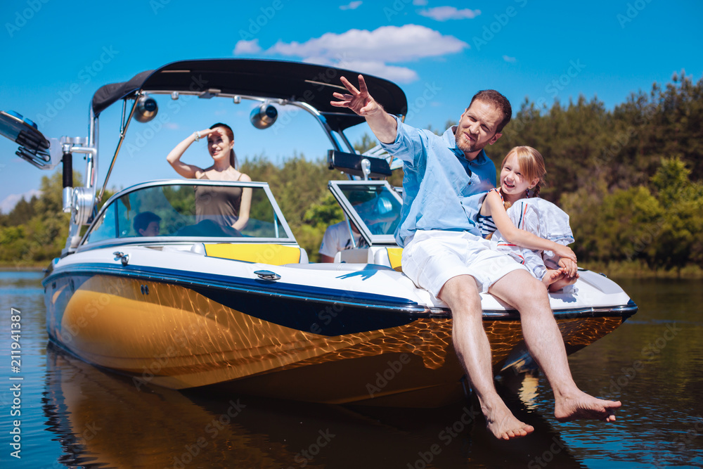 Wall mural Enjoying the travel. Loving young father pointing at riverbank and telling his daughter about their final destination while their family sailing the yacht