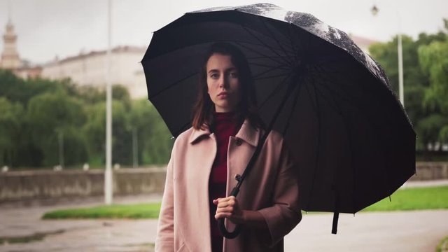 Young serious woman standing under umbrella in rain, sad weather in city