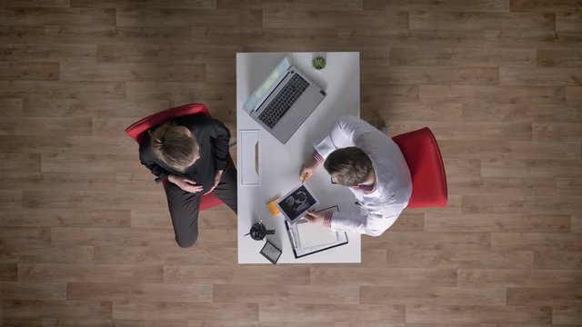 Doctor giving ultrasound to young pregnant woman, topshot, specialist advising future mother, sitting at table with laptop