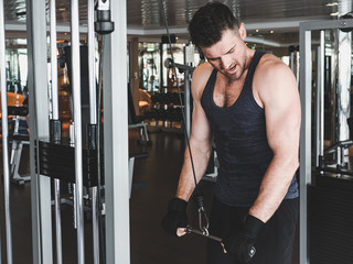 Stylish, muscular man in black training gloves and short sports shirt, performs strength exercises on sports equipment in an empty gym. Concept of a healthy lifestyle, strength and motivation