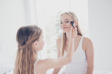 Obraz na płótnie Canvas Preteen schoolgirl style fun blonde long straight hair concept. Close up portrait of cute glad kid doing makeup for her older adopter sister applying maquillage on cheekbones holding brush in hand