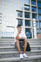 Fototapeta na wymiar Young attractive fro American businessman sitting on stairs in front of huge modern building and doing some work on laptop