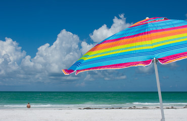 Tropical beach with colorful umbrella, blue sky, clouds, woman sitting alone.