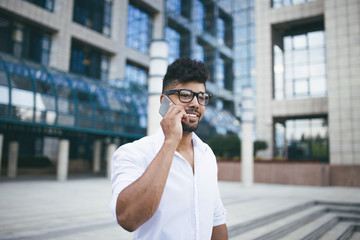 Positive and handsome Afro American man standing in front of huge modern business building smiling and talking on cell phone