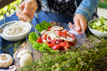 Woman hands adds chickpea hummus spread with sesame seeds tahini to vegetables salad with tomatoes and onion rings on big green salad leaves.  Raw vegan vegetarian food
