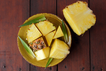 Pieces of pineapple on a yellow plate on wooden boards, pineapple with green leaves on a wooden background, salad of tropical fruits for breakfast, vegetarian food, copy space
