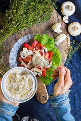 Woman hands adds chickpea hummus spread with sesame seeds tahini to vegetables salad with tomatoes and onion rings on big green salad leaves.  Raw vegan vegetarian food