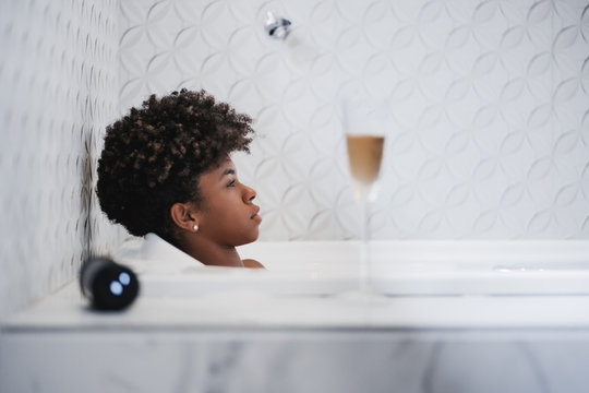 Side View Of A Tired Young Curly Black Girl Laying, Relaxing In The Bath Indoors And Listening To The Music Form A Wireless Speaker Next To Her And A Glass Of A White Wine In The Foreground