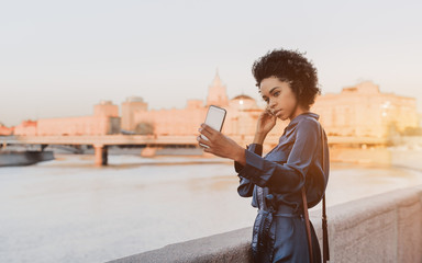 Dazzling young black tourist female near quay fencing is taking an evening selfie via smartphone; charming African-American girl in a blue dress is photographing herself on the cellphone near a river