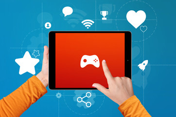 Boy's hand holding and touching the screen of tablet with game joystick on screen with award and achievement icons on blue background. Gamification concept