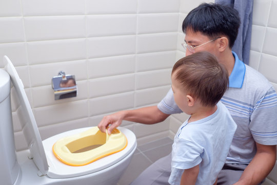 Father Training sleepy son to use toilet in bathroom, Cute little Asian 1 year old / 18 months toddler baby boy sitting on toilet with kid bathroom accessory, Potty / Toilet Training child concept