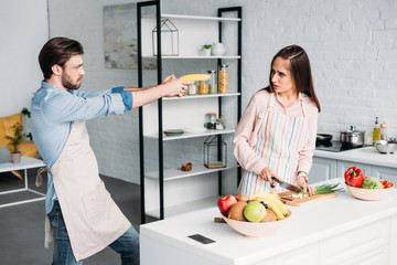 girlfriend cutting vegetables and serious boyfriend having fun with banana gun in kitchen
