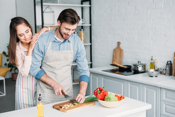 boyfriend cutting vegetables and girlfriend leaning on him in kitchen