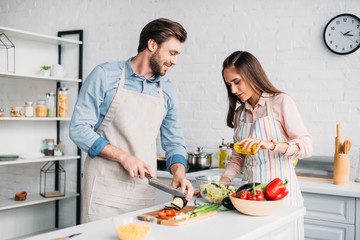boyfriend cutting vegetables and girlfriend pouring oil into salad in kitchen