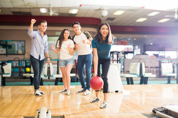 Friends Motivating Teen Girl Throwing Bowling Ball At Alley