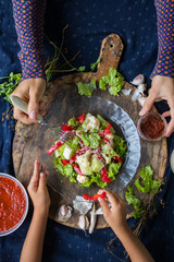 Woman hands and little girl hands holds fresh salad with raw cauliflower, red bell pepper, cucumber and dressed with white sesame seeds. Family lunch or dinner. Raw vegan vegetarian healthy food