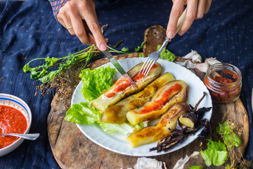 Woman hands cut roasted  grilled eggplants with fork and knife dressed with tomato garlic sauce, paprika, pepper and fresh salad leaves for lunch or dinner. Raw vegan vegetarian healthy food.